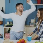 Attractive young joyful couple have fun dancing and singing while set the table for breakfast in the kitchen at home